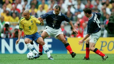 Getty Images Two football players in dark blue shirts challenge a player in a yellow shirt during a match.