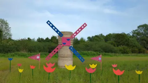 Comberton Young Farmers A windmill made from round straw bales with pink and blue sales and red, yellow pink and blue cut out flowers in front