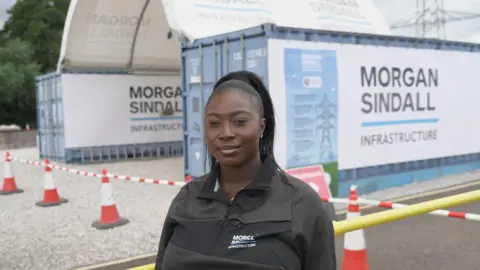 Hannah Karpel / BBC News A young woman in a black Morgan Sindall Infrastructure zip-up jacket standing in the foreground at a construction or infrastructure site. Behind the person are two blue site cabins or container units displaying large Morgan Sindall Infrastructure signs. Red-and-white traffic cones and barrier tape mark off a gravel area, with a yellow safety barrier running across the lower right of the image. Additional site signage is attached to one container, and electricity pylons are visible in the background.