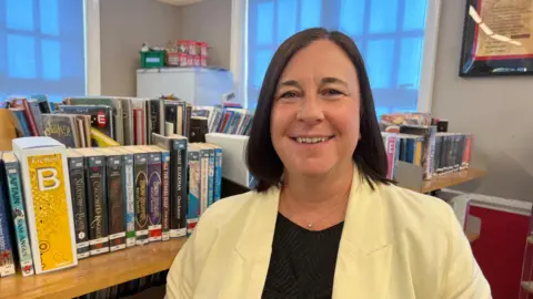 Laura Mackie is pictured wearing a yellow blazer-style jacket with a black top underneath. She is smiling widely at the camera. She has dark brown, shoulder-length hair.
Behind her is a book shelf filled with books. 