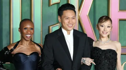 EPA Cynthia Erivo, Jon M. Chu and Ariana Grande pose together at the UK premiere. They link arms and smile. Cynthia Erivo and Ariana Grande wear black gowns while Jon M. Chu wears a smart black suit jacket over a white shirt and tie.