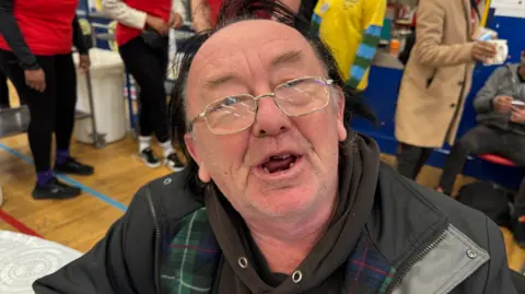 A man with glasses and dark hair is prominent in the photo. He is wearing a dark coat and other people in the background are blurred.