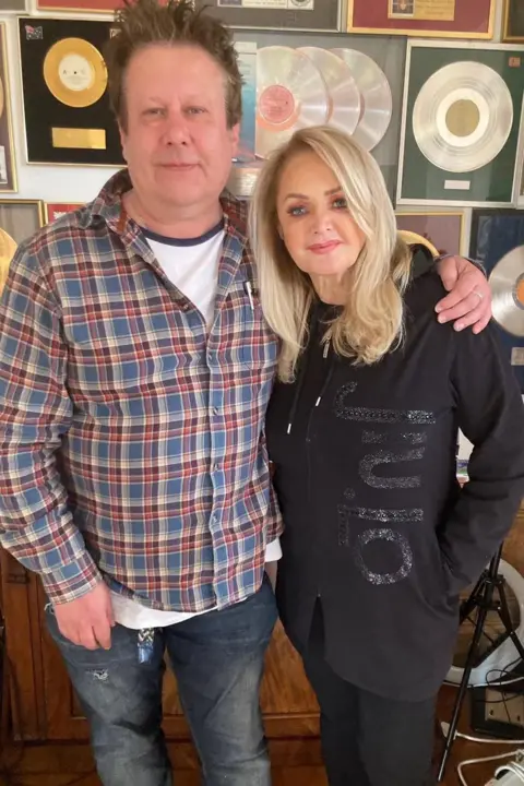 Rob Marshall A man with spiky brown hair wearing a red and blue check shirt and jeans, with his arm around a glamorous blonde woman wearing all black. Behind them is a wall with lots of framed records on it.