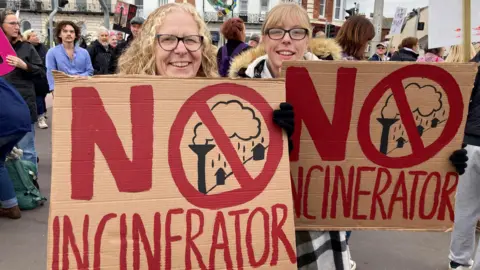 Two women with blonde hair and glasses holding up placards reading "no incinerator", with a crowd of people standing behind them.