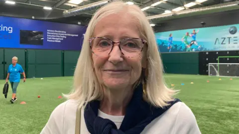 Pip Loinger has shoulder length blonde hair and is wearing glasses and a cream jumper. In the background are the sports pitches.