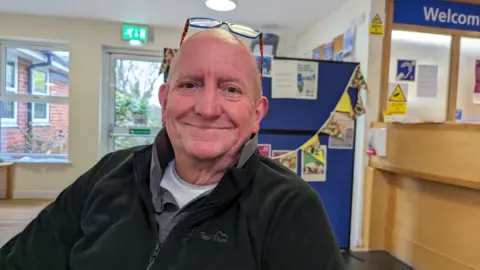 Philip Ratsey sitting in a doctors' waiting room. He is wearing a black fleece and glasses pushed onto his head and he is smiling. 