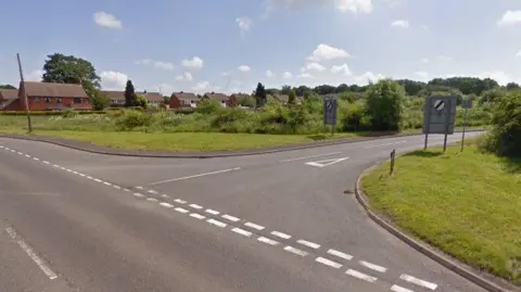 A road junction, with houses in the background. It is a semi-rural setting, with trees and green space visible. There are road signs indicating the road is set to the national speed limit.