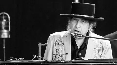 Getty Images Bob Dylan, wearing a black hat and a light jacket, is seen performing on stage