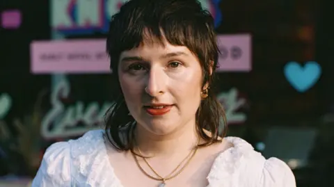 Theodore van Duin A brunette young-woman with soft facial features and brown eyes. She has pale skin, and coral-toned lips, and her hair is styled in a wolf-cut, which is a combination of the 1970s shag and the modern mullet. She is wearing a white, frilly v-neck top and has two gold necklaces on.