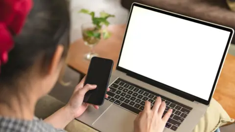 Woman types on computer while holding an iPhone.