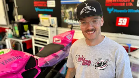 BBC Blake Kelly wears a grey t-shirt, clear spectacles, and a dark cap. He smiles. He sits next to a hot pink motorcycle.