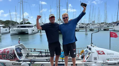 ATLANTIC DASH Tim Foster, a man with a cap and navy shirt on the left, and Mike Reed, a ginger man with a blue long-sleeve top, stand with their arms up to celebrate front of the small white boat with a Manx flag.