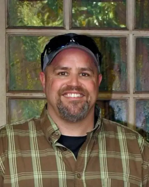 Luke Thompson Luke Thompson, with a goatee beard, smiling and wearing a brown and green checked shirt and a baseball cap.