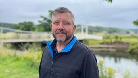 Mark Ryan wearing a dark coat, standing on a riverbank 