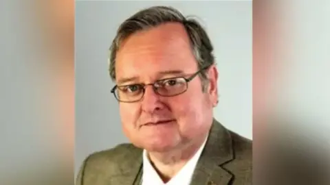 Warwickshire County Council Councillor Edward Harris wears glassed and a brown suit jacket.