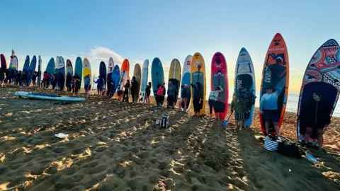 Paddleboarders lined up with their boards on a sandy beach.