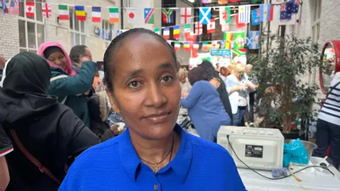 BBC Soliana Rama stands in front of a room covered in flags from across the world. There is a crowd behind her and people are queuing for food at multiple tables