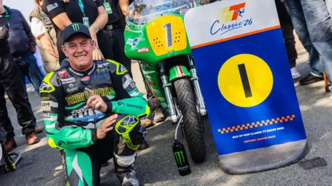 CLASSIC TT John McGuinness, in green and black Team Winfield leathers, kneeling next to the bike and a board with the number one on it.