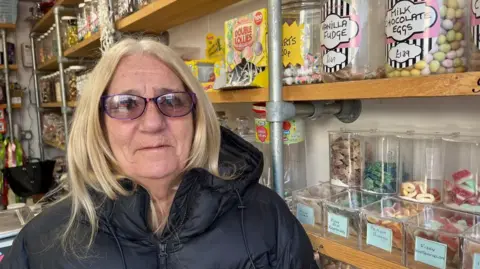 Andrew Turner/BBC Yasmin Harwood, has blonde below-the-shoulder length hair and is wearing a black coat and spectacles. She is standing next to shelves of jars of sweets.