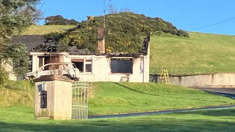 The remains of a house after a fire. The roof is mostly gone. The car has also been burnt out. Green fields are surrounding the property. 