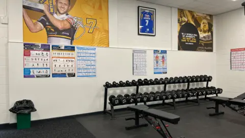 Michelle Lyons The interior of the Rob Burrow Fitness Suite. There are weights racked and a weight bench, as well as posters of Burrow on the wall and a Leeds Rhinos shirt.