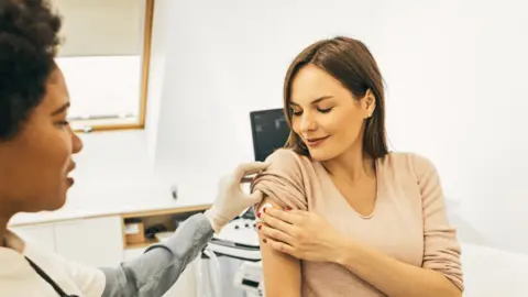 A doctor and a patient after a vaccine. The female patient is holding a cotton ball on her upper arm and looking down at it. The doctor is wearing latex gloves and holding the patient's sleeve up. 