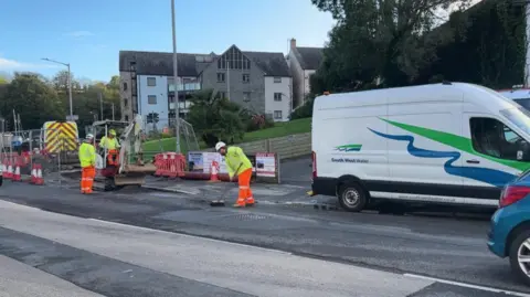 A man wearing a yellow hi vis jacket and orange high vis trousers sweeps water off the street and into the gutter. Behind him is a South West Water van parked half on the pavement. In front of him two more workers are operating a small digger which is surrounded by cones and fencing.