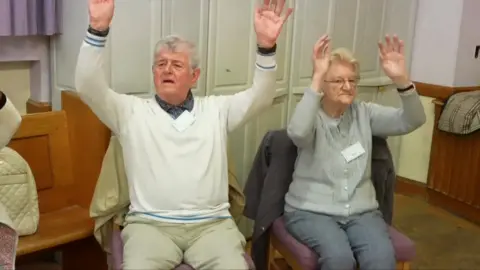 A man and a woman sitting on chairs with their hands raised in the air with their palms forward.