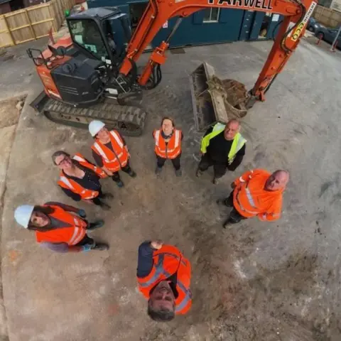 Context One An aerial selfie of seven people wearing orange hi-viz and hard hats looking up at the camera as they stand in front of a mechanical digger