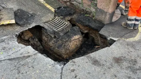 A sinkhole in a road nearby to a drain. There is a wall in the background of the picture and some feet in orange high vis trousers.