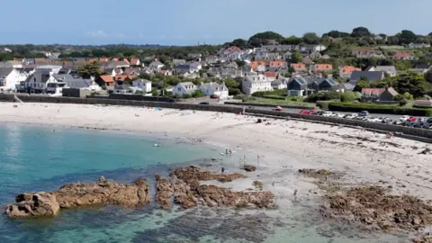BBC Cobo Bay in Guernsey during the summer. White sand stretches along the coast road. people are in the turquoise water. A rock formation in the sea is surrounded by seaweed. Behind the road are white houses with terracotta roofs.
