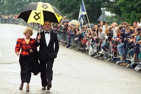 Getty Images Alex Salmond pictured with his wife Moira at the Braveheart premiere in Stirling in 1995. Moira wears a tartan blazer while Alex wears a black suit jacket, white shirt and black bowtie. He is carrying a black and yellow umbrella.