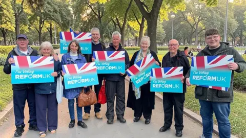 An image posted by Reform UK which appears to have been modified to change the background behind a group of campaigners holding Reform UK signs. Behind them are trees and a park with grass and bushes. They are each smiling and holding up pale blue signs with a section of union jack flag on them, bearing the words "I'm voting Reform UK".