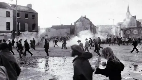 Getty Images A black and white image shows demonstrators running from tear gas. There are streets in the background, and the smoke from the tear gas can be seen rising from different parts of the ground.