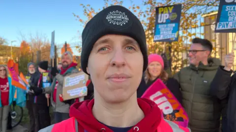 A woman in a beanie hat with National Education Union on in front of a picket line with people holding banners