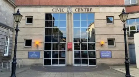 The Civic Centre in Stoke, a light-coloured brick building with large panelled windows which are about two-storeys in height. There are two traditional-style lampposts in the foreground.
