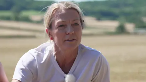 A head-and-shoulders picture of Zoe Tucker in a white t-shirt in a field, with hedges and trees in the background 