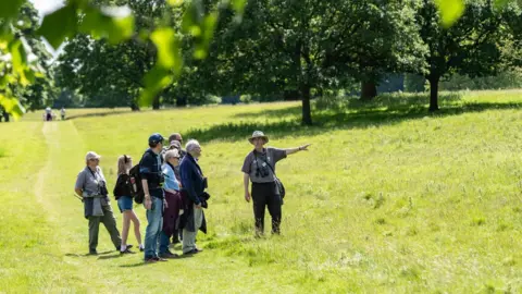 A sunny field with trees in the background and a group of walkers standing around a man who is pointing into the distance and wearing binoculars around his neck.