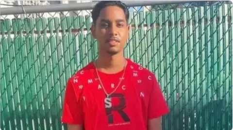 Family  Shawn Seesahai wearing a red t-shirt and chain around his neck, standing against a green fence