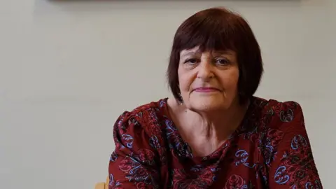 Stephen Huntley/BBC Hazel Simmons, looking straight at the camera, with short bobbed dark hair, wearing a burgundy and blue and pink patterned dress. 