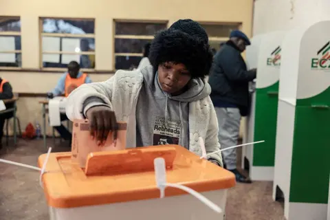 A woman in a black woolly hat and thick white cardigan places a ballot paper in a plastic ballot box with an orange lid. Some polling booths and election officials can be seen behind her. 