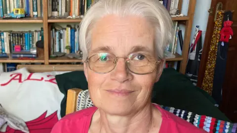 A woman with short white hair smiles at the camera. She is wearing glasses and a pink top. In the background is rows of bookshelves and a sofa throw featuring the Welsh flag.