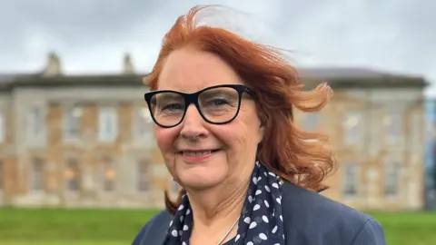 BBC Anne Robson smiles at the camera. She has shoulder length red hair and black-framed glasses. She is wearing a navy suit jacket with navy scarf decorated with white dots. Behind her is a large Georgian-style manor house.