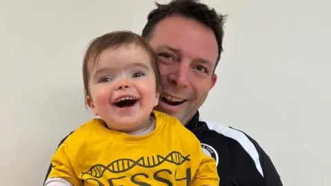 A man in a black tracksuit top holds up his young daughter who is wearing a yellow T shirt and is smiling broadly 