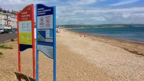 Beach safety signs at the top of the beach at Greenhill, with a street to the left and blue sea and coastal hills in the background