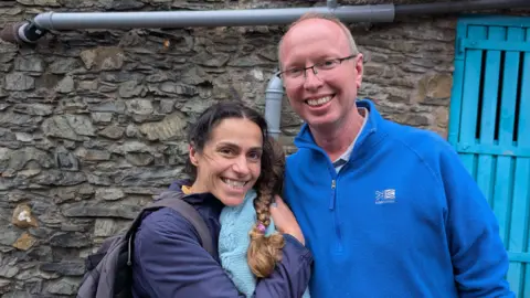 Stuart and Sufia Mills are standing side by side, holding a small girl, who is looking away from the camera. Sufia has dark hair tied back and is wearing a blue coat and carrying a rucksack. Stuart has fair receding hair and wears glasses and a bright blue sweater. They are smiling at the camera. The girl is wearing a blue jumper and has her brown hair in plait. They are standing in front of a stone wall with a blue gate.