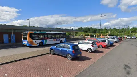 Google The large car park at Priory Park and Ride. Cars are parked in bays while a single-decker bus waits outside the single-storey bus station. A row of green trees can be seen in the background.