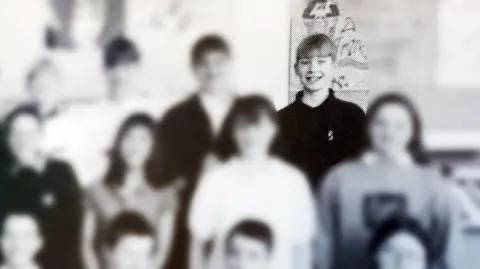 An old school photograph in black and white. It has been blurred so only the face of a smiling teenager is visible at the back of the class. 