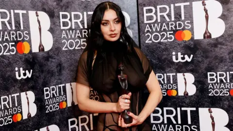 Getty Images Charli XCX in her sheer dress holding a Brit Award in front of a Brits-branded backdrop