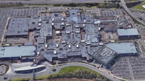 Aerial view of a large shopping centre. There multiple buildings joined together and large car parks around.